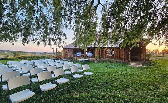Outdoor wedding ceremony setup with white chairs on green lawn beside rustic wooden barn.