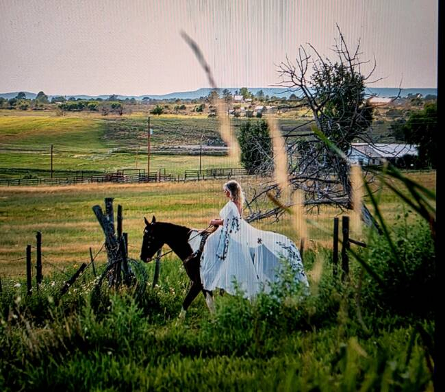 Bride in flowing white dress riding horse through scenic countryside field at rustic ranch wedding venue.