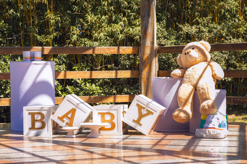 Outdoor baby shower setup featuring decorative BABY blocks, teddy bear, and gift table in a rustic garden setting.