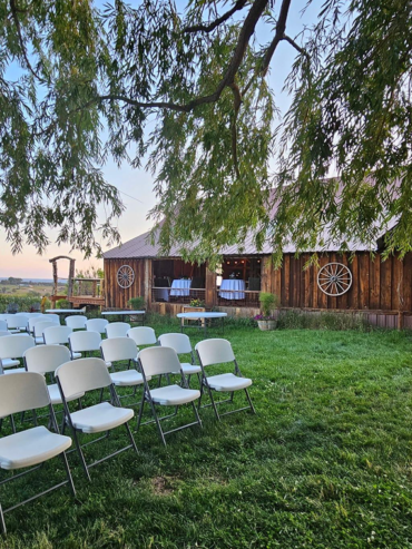 Outdoor wedding ceremony setup with white chairs on green lawn beside rustic wooden barn.