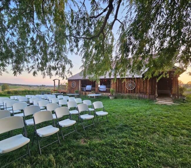 Outdoor wedding ceremony setup with white chairs on green lawn beside rustic wooden barn.