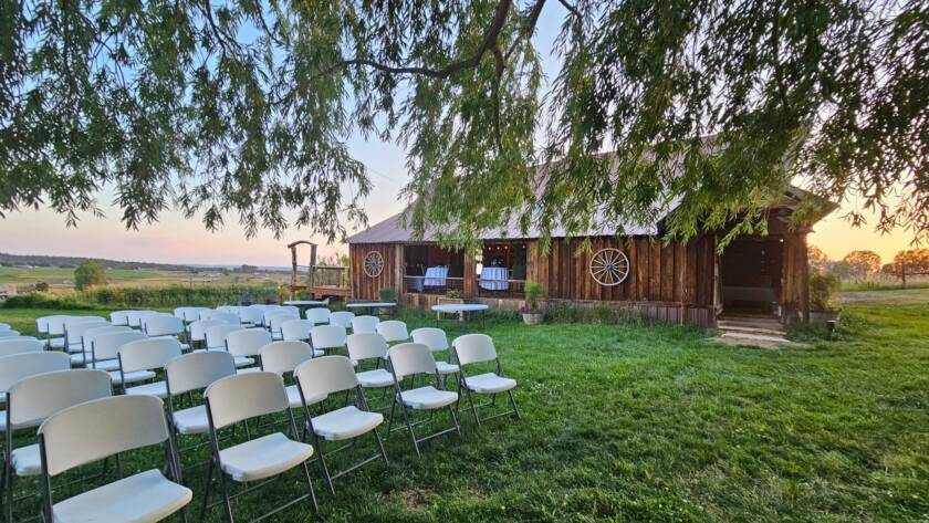 Outdoor wedding ceremony setup with white chairs on green lawn beside rustic wooden barn.