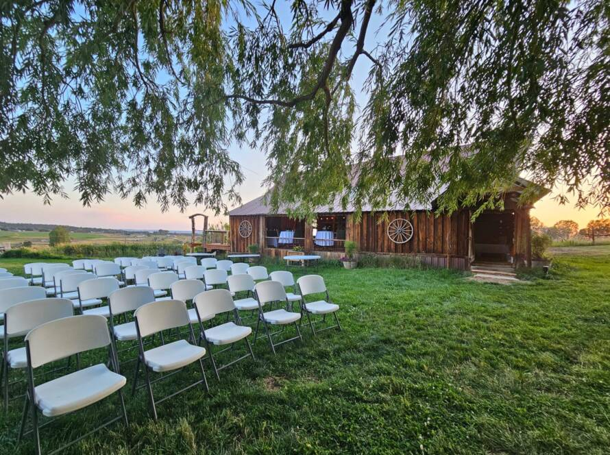Outdoor wedding ceremony setup with white chairs on green lawn beside rustic wooden barn.