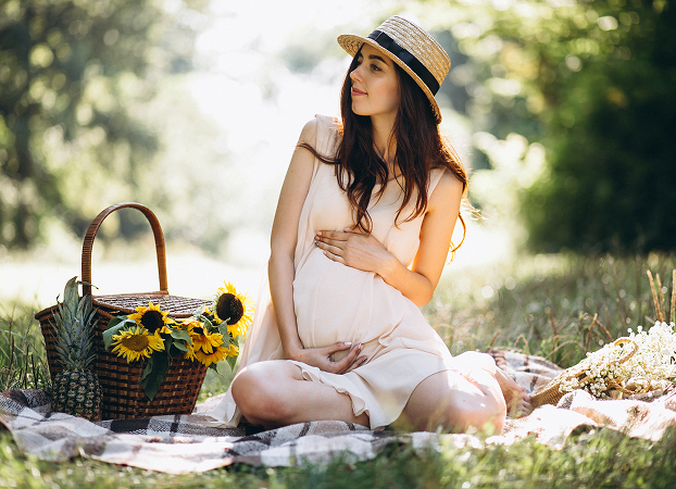 Expecting mother sitting outdoors on picnic blanket with sunflowers, celebrating pregnancy in a natural garden setting.