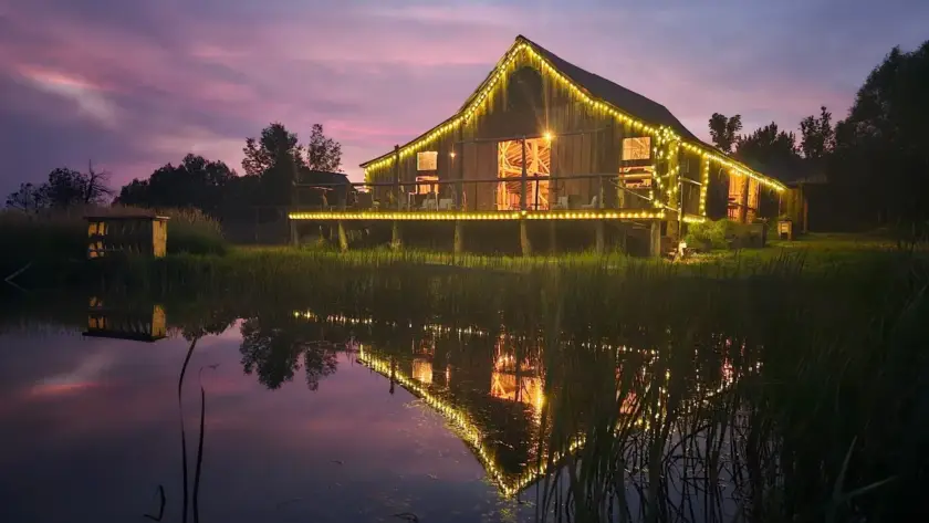 Rustic barn at Three Willows Ranch glowing with evening lights reflected on the water in Colorado