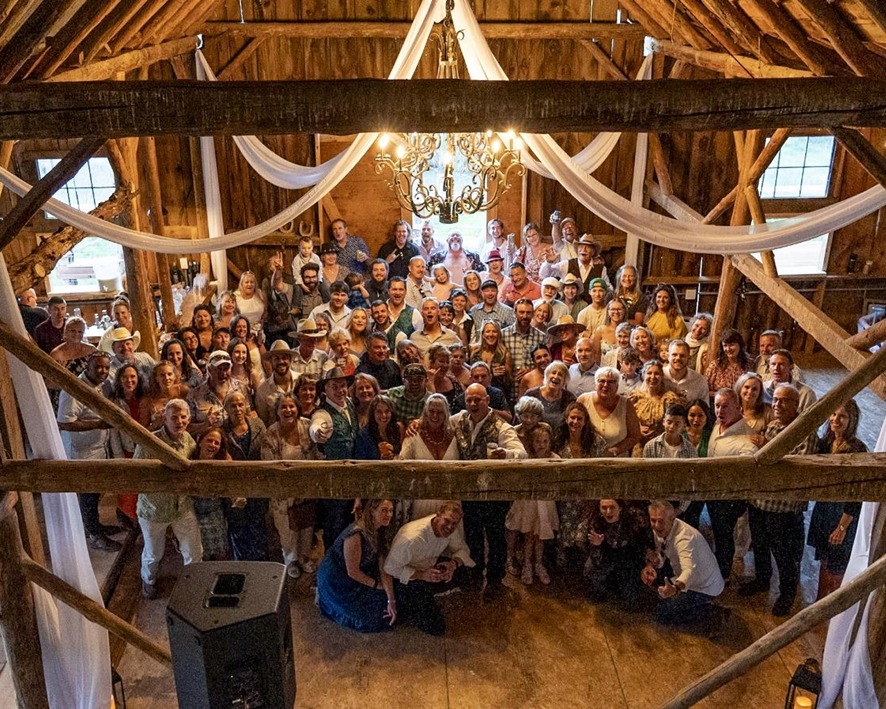 Wedding guests gathered inside a historic wooden barn reception space with chandelier and draped fabric at a rustic Colorado ranch venue.