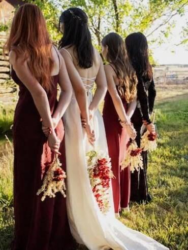 Bride and bridesmaids standing outdoors at a rustic Colorado ranch wedding, holding bouquets behind their backs beside a barn and trees.