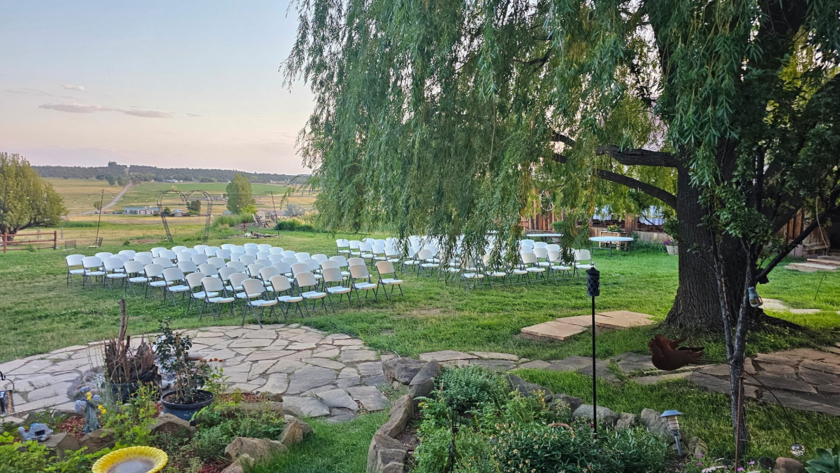 Outdoor wedding ceremony setup with white chairs on green lawn beneath large willow tree at rustic ranch venue.