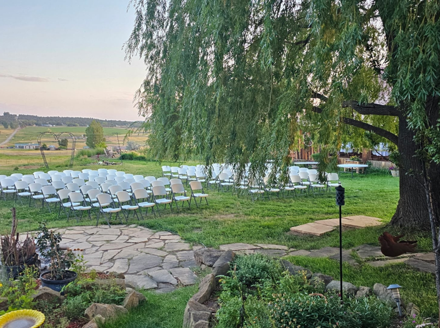 Outdoor wedding ceremony setup with white chairs on green lawn beneath large willow tree at rustic ranch venue.
