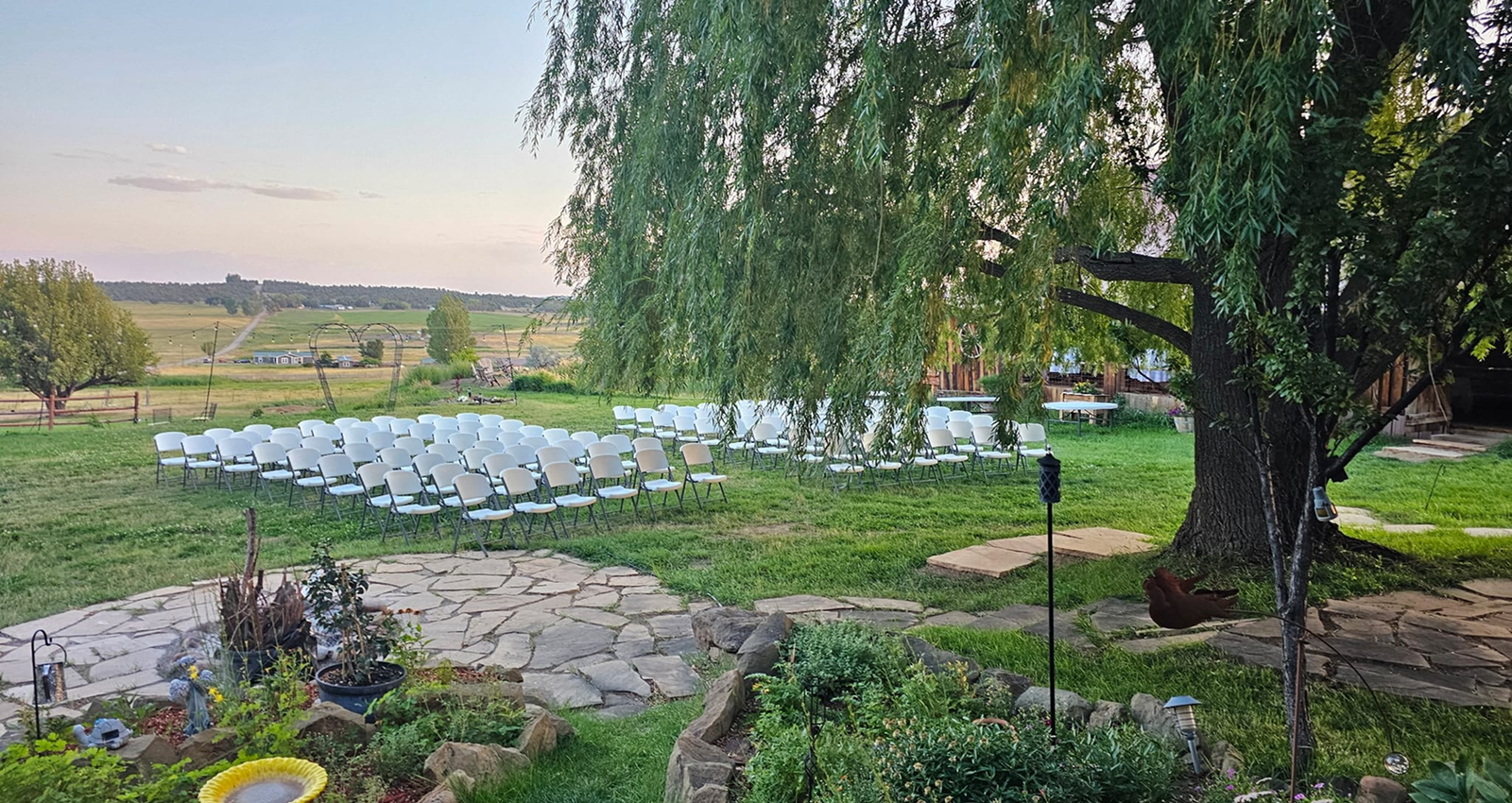 Outdoor wedding ceremony setup with white chairs on green lawn beneath large willow tree at rustic ranch venue.