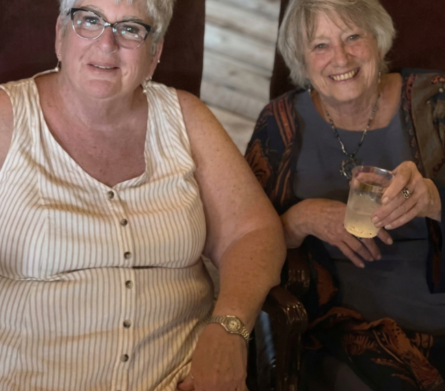 Two women smiling and relaxing with drinks while seated inside a rustic barn during a warm and welcoming ranch event.