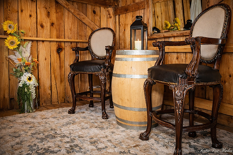 Rustic wooden barn seating area with vintage chairs, a wooden barrel table, lantern decor, and sunflower floral arrangement.