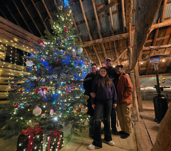 Decorated Christmas tree with colorful lights inside a rustic wooden barn with people standing beside it during a holiday gathering.