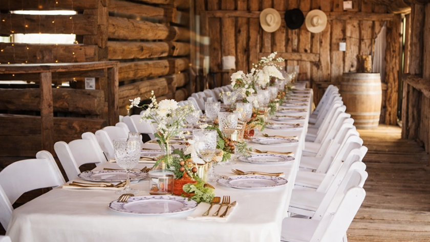 Rustic barn banquet table with white chairs, elegant place settings, and floral centerpieces.