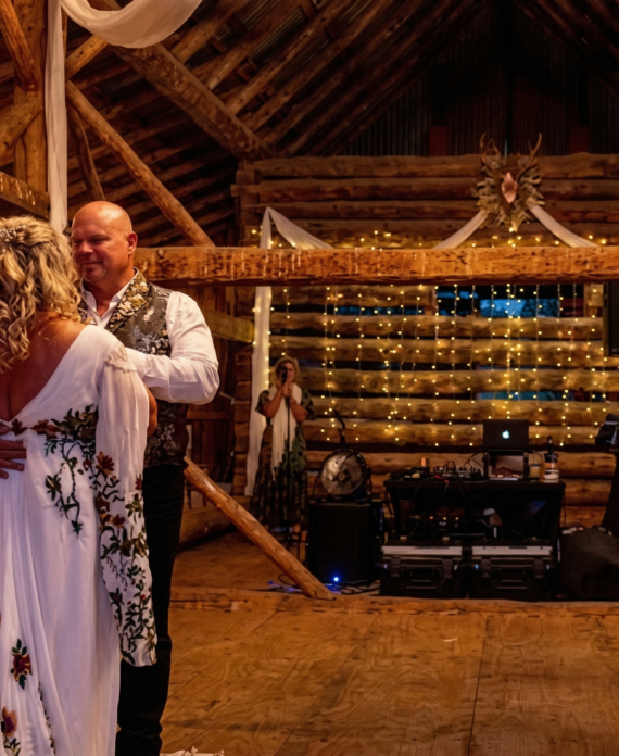 Bride and groom dance on a wooden dance floor inside a warmly lit rustic barn wedding venue.