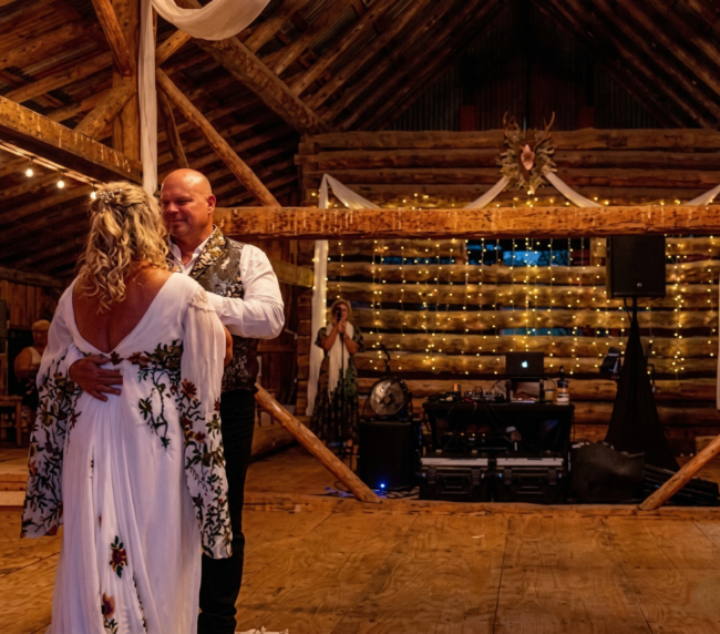 Bride and groom dance on a wooden dance floor inside a warmly lit rustic barn wedding venue.