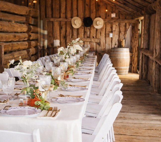 Long decorated dining table with white chairs and floral centerpieces inside a rustic wooden barn wedding reception venue.