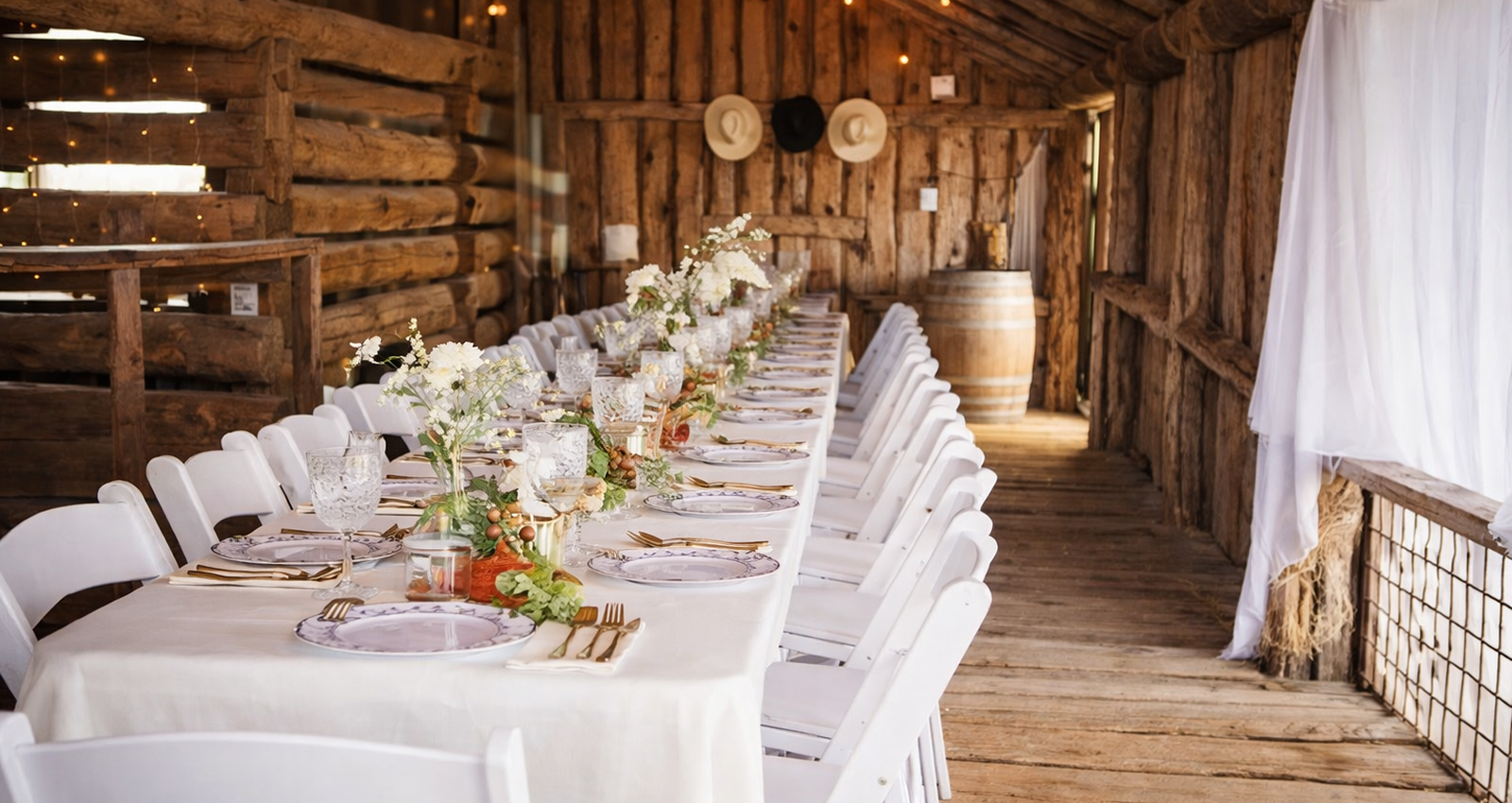 Long decorated dining table with white chairs and floral centerpieces inside a rustic wooden barn wedding reception venue.