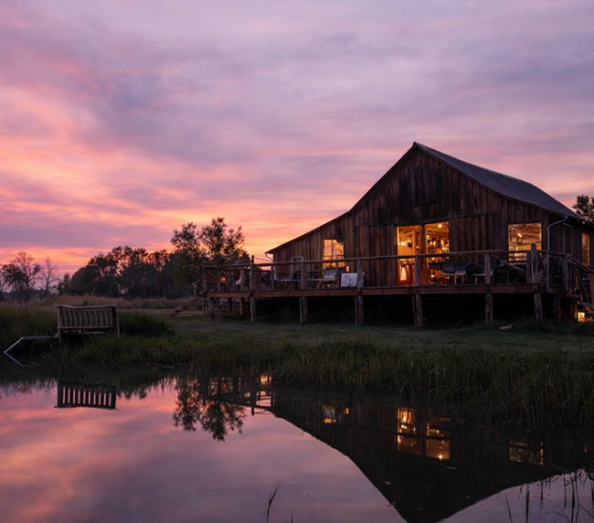 Rustic wooden ranch barn beside a calm lake at sunset with warm lights reflecting on the water and colorful evening sky.