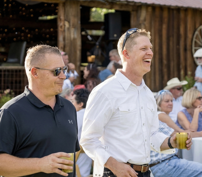 Guests enjoying drinks and laughing during an outdoor cocktail hour in front of a rustic barn at a Colorado ranch gathering.