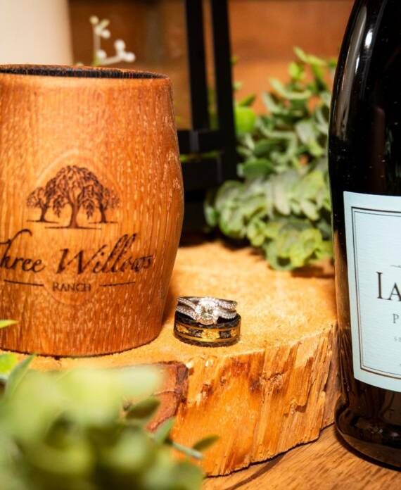 Wedding rings beside a Three Willows Ranch wooden cup and prosecco bottle on a natural wood slice, capturing elegant rustic wedding details.