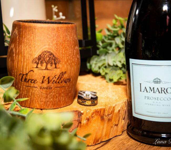Wedding rings beside a Three Willows Ranch wooden cup and prosecco bottle on a natural wood slice, capturing elegant rustic wedding details.