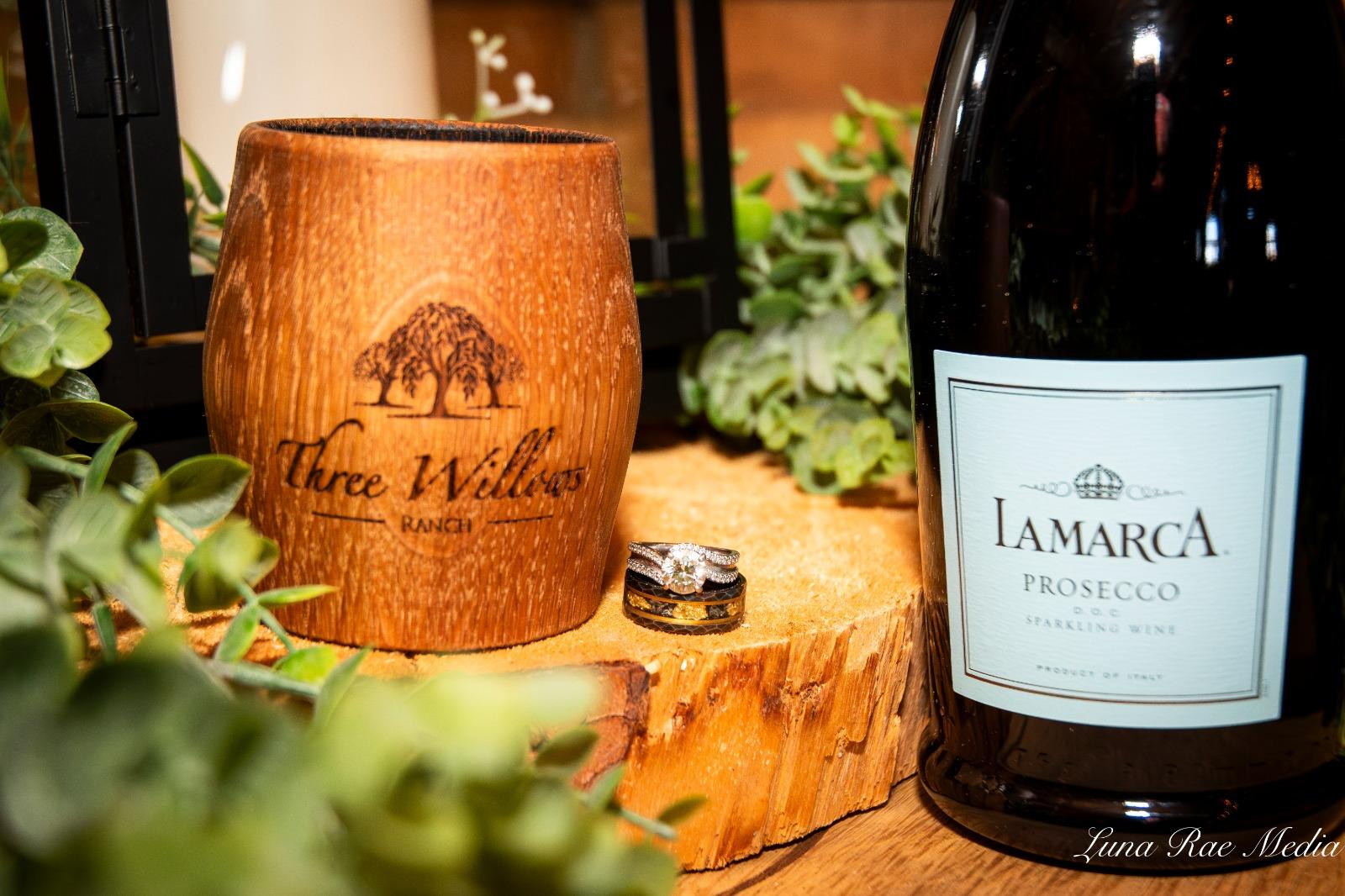 Wedding rings beside a Three Willows Ranch wooden cup and prosecco bottle on a natural wood slice, capturing elegant rustic wedding details.