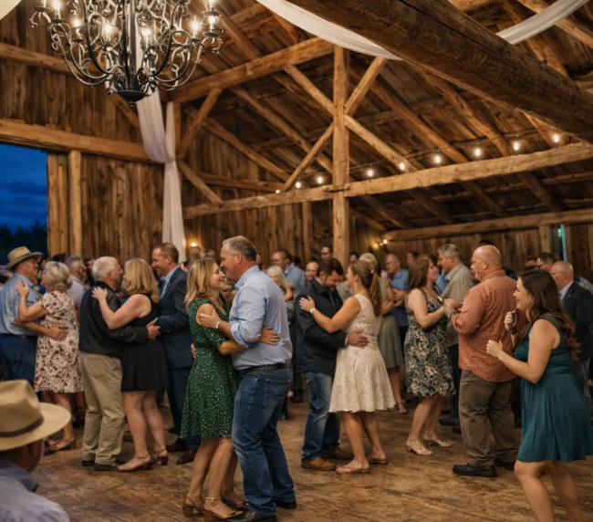 Rustic barn wedding reception with couples dancing under chandelier lighting and wooden beams in a warm, cozy indoor venue