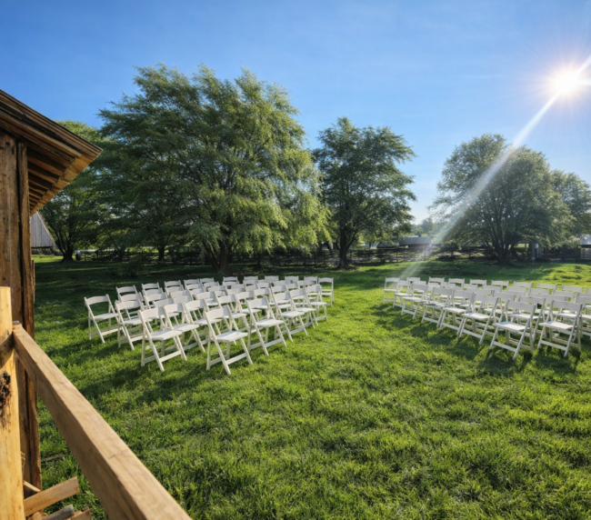 Outdoor rustic wedding ceremony setup with white folding chairs arranged on green grass under bright sunlight beside a wooden barn and trees