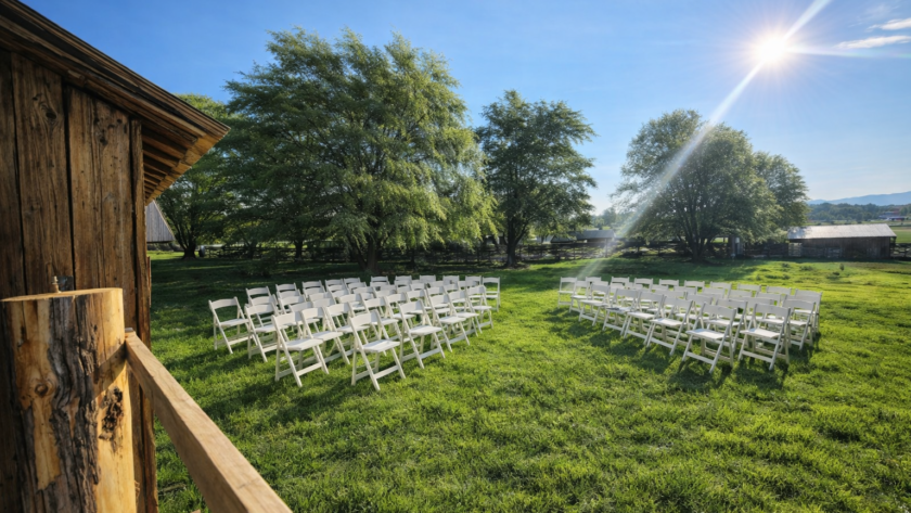 Outdoor rustic wedding ceremony setup with white folding chairs arranged on green grass under bright sunlight beside a wooden barn and trees
