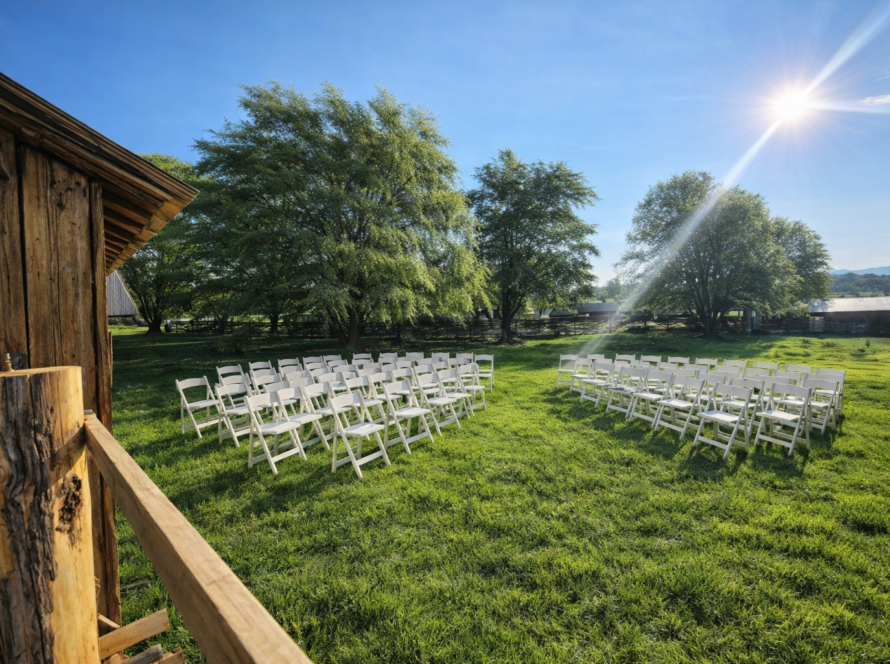 Outdoor rustic wedding ceremony setup with white folding chairs arranged on green grass under bright sunlight beside a wooden barn and trees