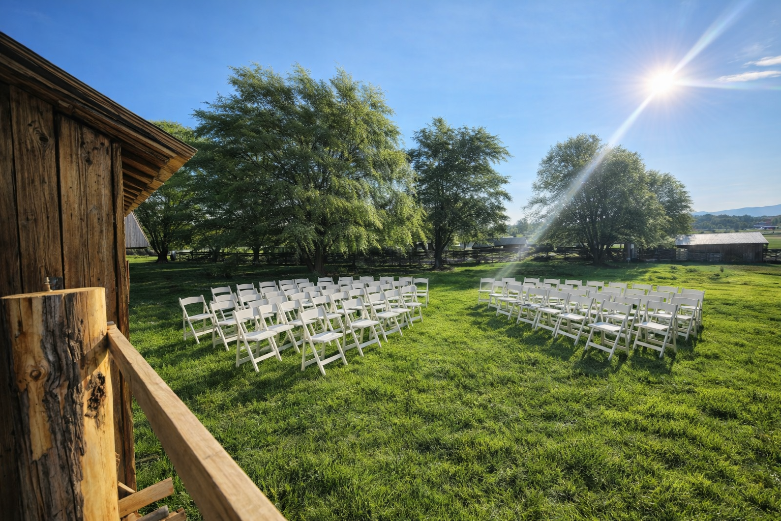 Outdoor rustic wedding ceremony setup with white folding chairs arranged on green grass under bright sunlight beside a wooden barn and trees