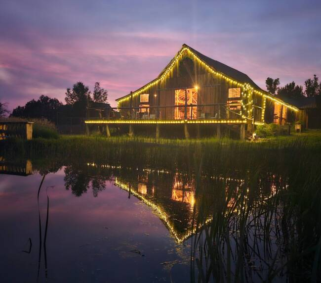 Rustic barn at Three Willows Ranch glowing with evening lights reflected on the water in Colorado