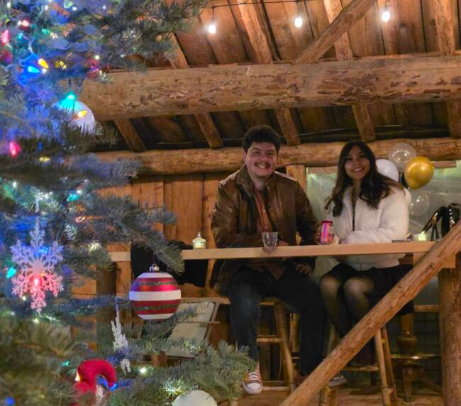 Couple enjoying a festive holiday celebration inside a rustic barn with a decorated Christmas tree and warm string lights at Three Willows Ranch.