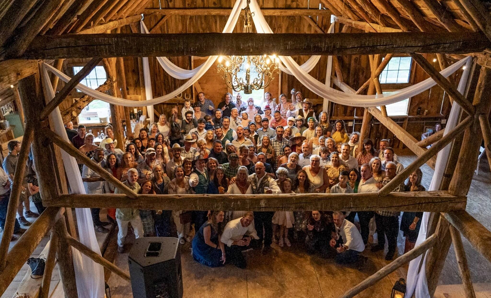 Wedding guests gathered inside a historic wooden barn reception space with chandelier and draped fabric at a rustic Colorado ranch venue.