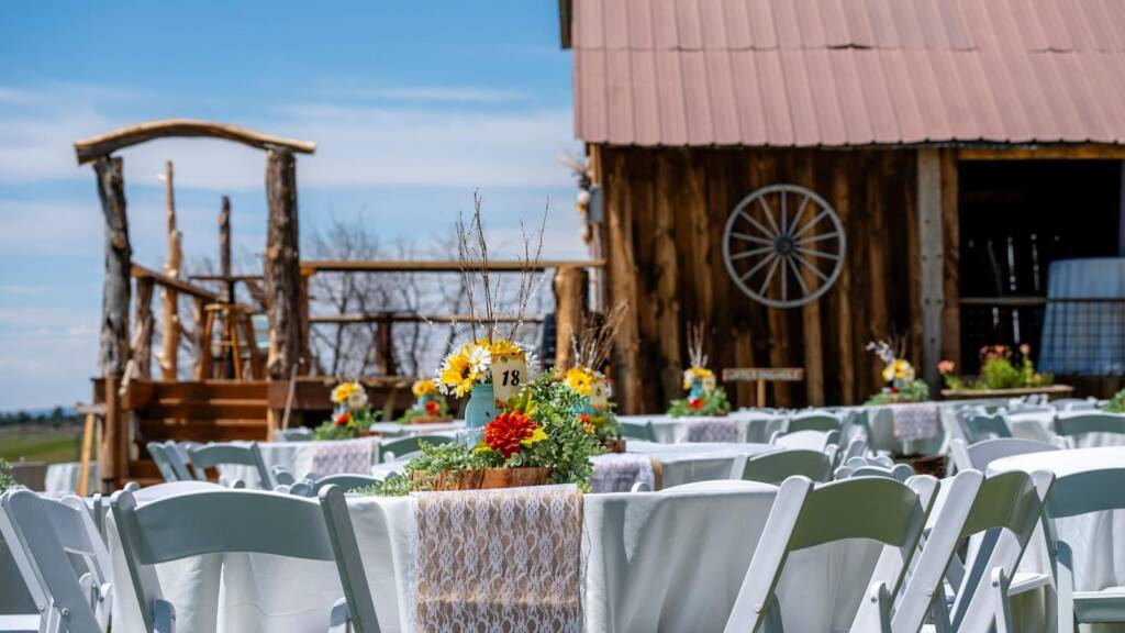 Long reception table with white chairs, floral decor, and string lights inside rustic barn wedding venue
