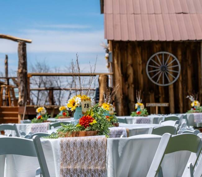 Long reception table with white chairs, floral decor, and string lights inside rustic barn wedding venue