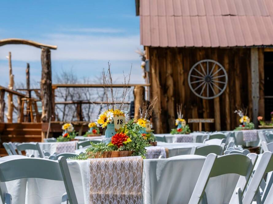Long reception table with white chairs, floral decor, and string lights inside rustic barn wedding venue