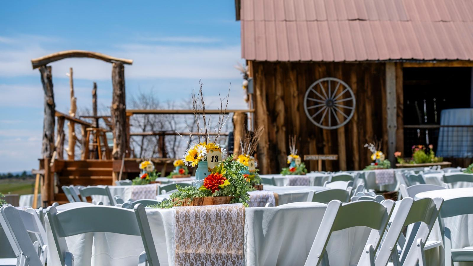 Long reception table with white chairs, floral decor, and string lights inside rustic barn wedding venue