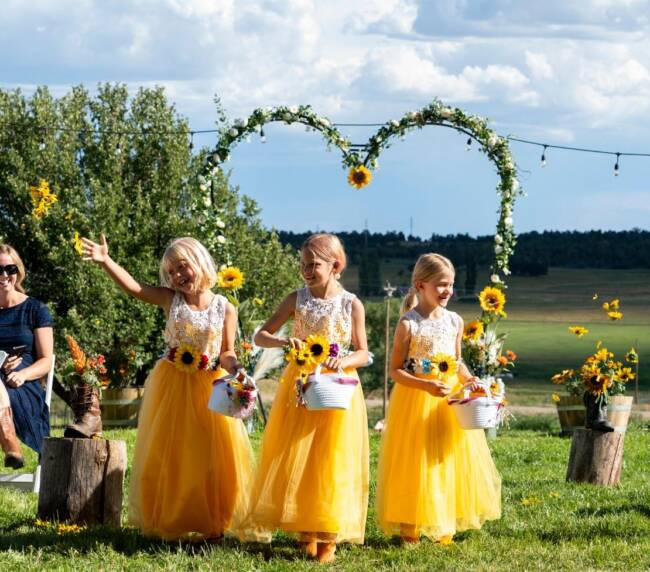 Flower girls in yellow dresses tossing petals during an outdoor wedding ceremony with a sunflower-decorated arch and scenic countryside at Three Willows Ranch in Colorado.