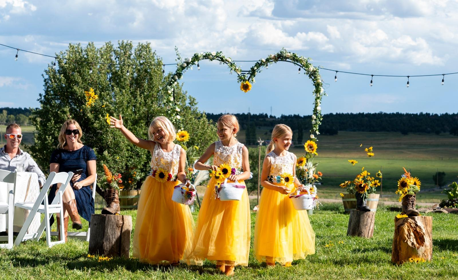 Flower girls in yellow dresses tossing petals during an outdoor wedding ceremony with a sunflower-decorated arch and scenic countryside at Three Willows Ranch in Colorado.