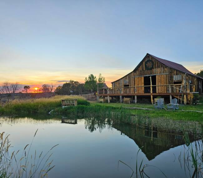 Wooden ranch barn at sunset reflecting over peaceful pond with countryside landscape view.