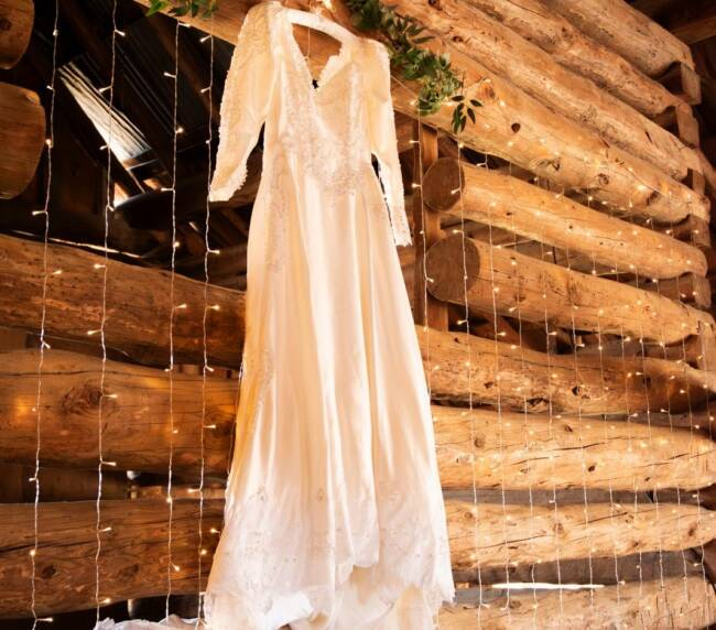 Elegant lace wedding dress hanging on rustic wooden barn wall with greenery and warm string lights