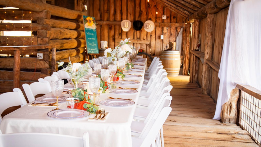 Rustic barn wedding reception setup with long decorated table, white chairs, floral centerpieces, and warm string lights at Three Willows Ranch in Colorado.