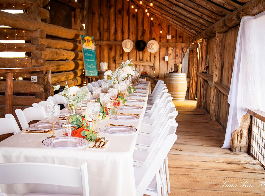 Rustic barn wedding reception setup with long decorated table, white chairs, floral centerpieces, and warm string lights at Three Willows Ranch in Colorado.