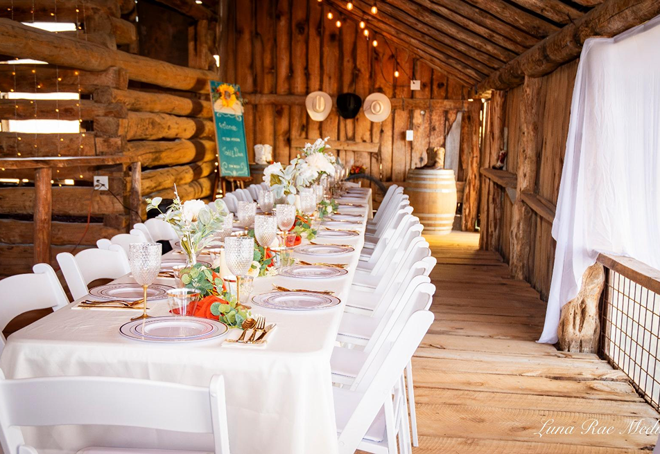 Rustic barn wedding reception setup with long decorated table, white chairs, floral centerpieces, and warm string lights at Three Willows Ranch in Colorado.
