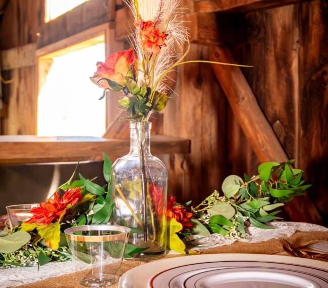 Rustic wedding table centerpiece with flowers and glass bottle near sunlit barn window