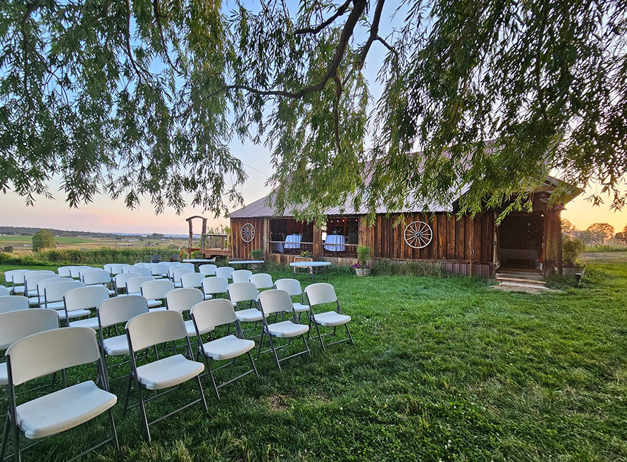 Outdoor wedding ceremony setup with white chairs on green lawn beside rustic wooden barn.