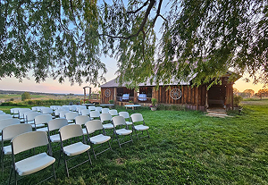 Outdoor wedding ceremony setup with white chairs on green lawn beside rustic wooden barn.