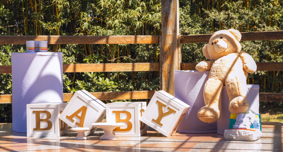 Outdoor baby shower setup featuring decorative BABY blocks, teddy bear, and gift table in a rustic garden setting.